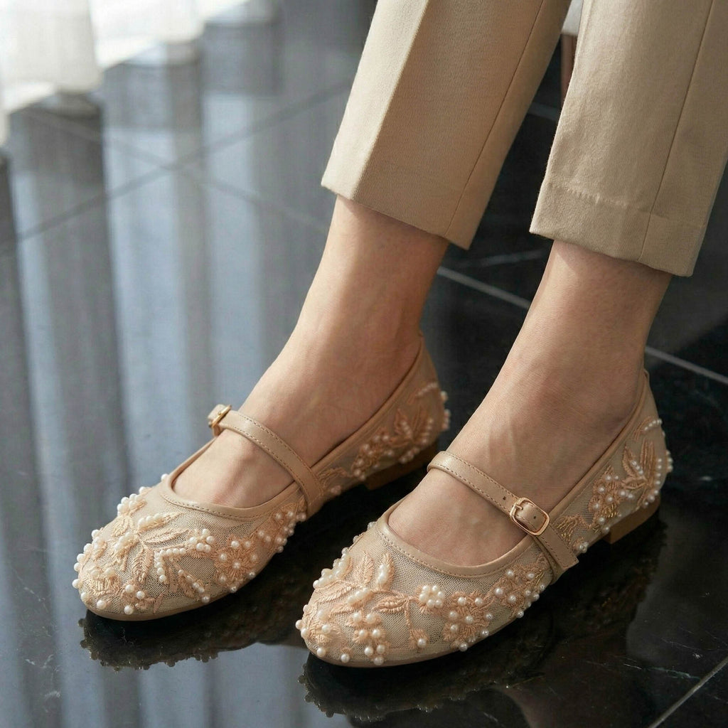 Beige flats with floral embroidery and pearl details worn by a person on a reflective surface.