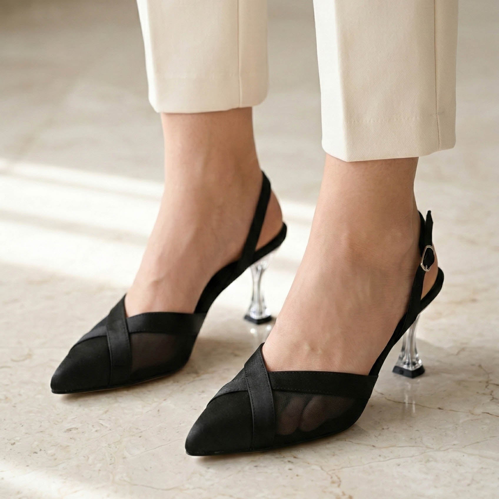 Black pointed-toe shoes with slingback straps worn with beige pants on a light wooden floor.