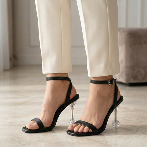 Black high-heeled sandals worn with white pants on a light wooden floor.