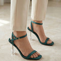 Teal high-heeled sandals worn with beige pants on a light wooden floor.