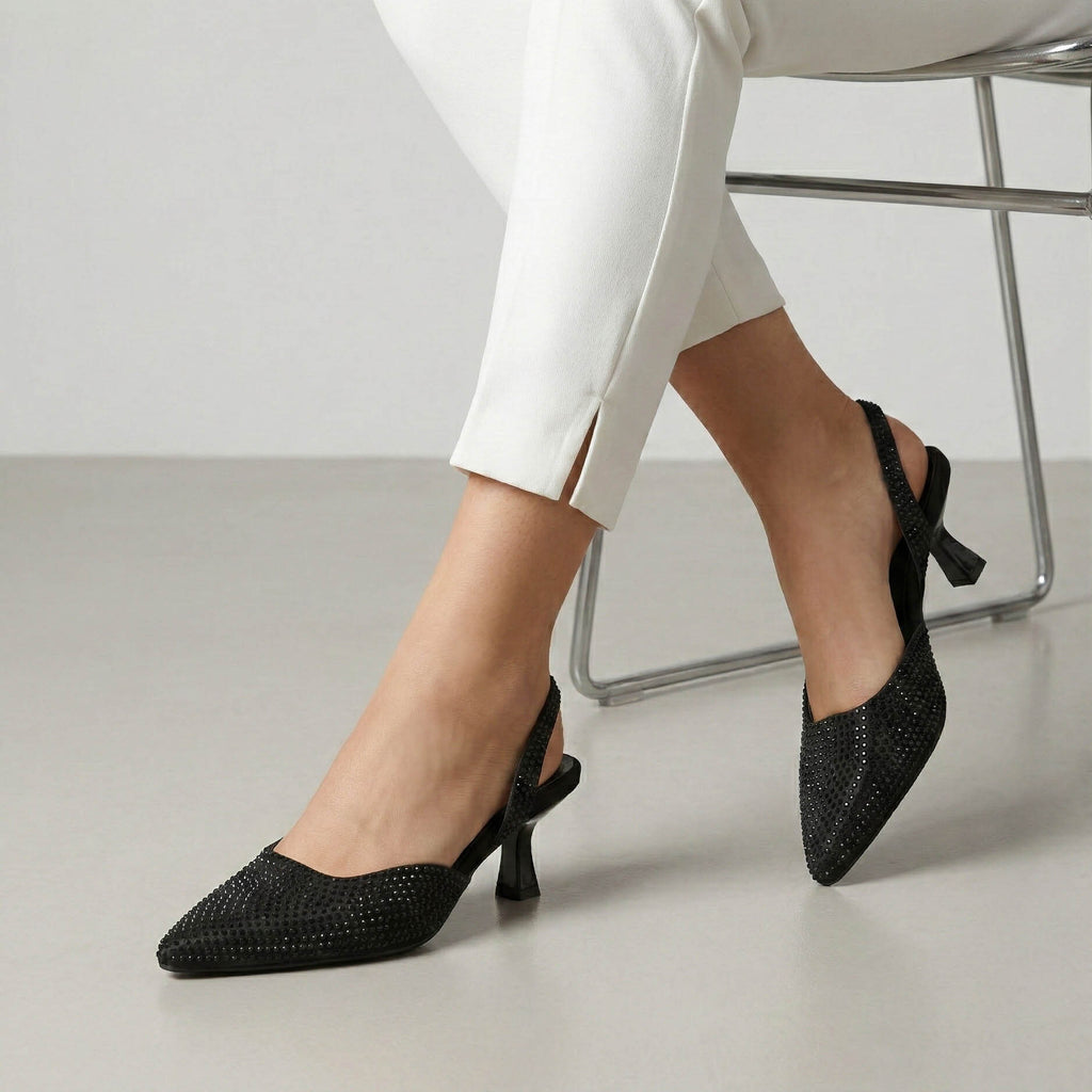 Black pointed-toe shoes with rhinestone details worn by a person sitting on a chair.