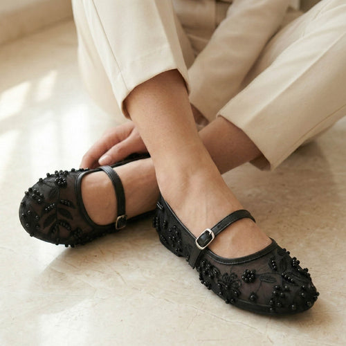Black embellished shoes worn with beige pants on a light wooden floor.